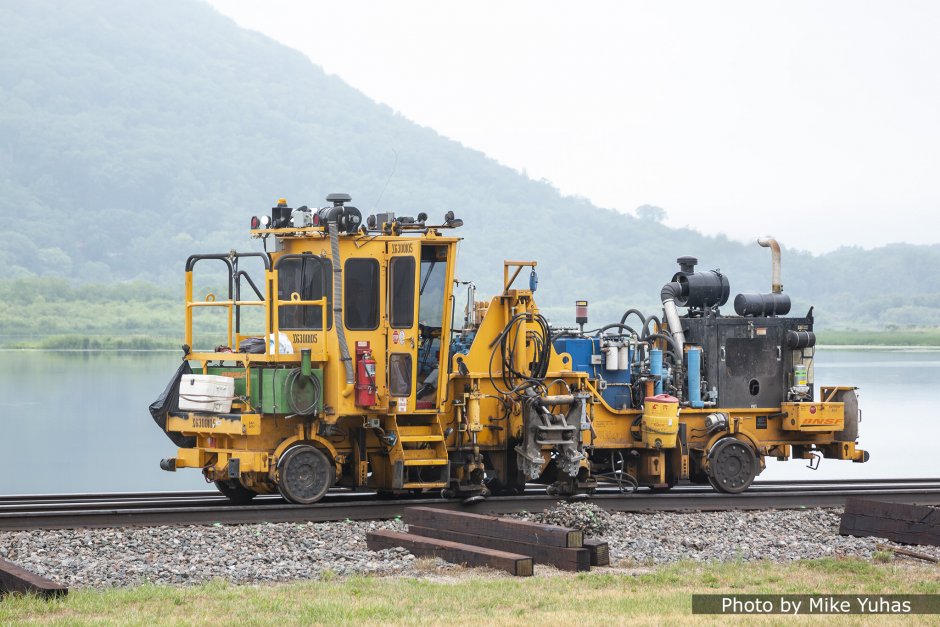 The first tie remover pulls the old ties partway out from beneath the rail. In this step, the rails are lifted so the tie (and tie plate) may be easily slid out. The bulging pile of ballast hints at the amount of force this machine exerts on the tie.