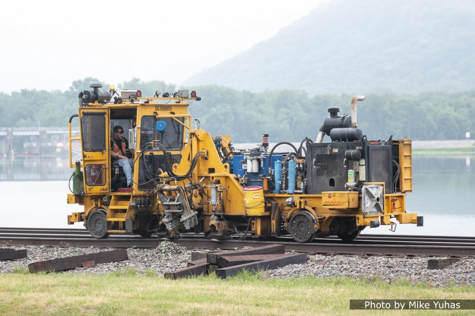 The first tie remover pulls the old ties partway out from beneath the rail. In this step, the rails are lifted so the tie (and tie plate) may be easily slid out. The bulging pile of ballast hints at the amount of force this machine exerts on the tie.