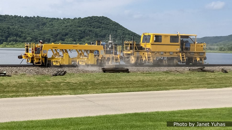 After the weekend, work resumed on Monday morning, June 28 with another multi-function machine. The first unit, a double broom, sweeps ballast off the tops of the ties. The second unit is a track stabilizer. Following tamping work or ballast bed cleaning, not all ballast stones are lying in a consolidated and stable position. This machine applies a horizontal vibration, crosswise to the track. This is transmitted to the ballast bed achieving a rearrangement of the ballast stones to produce a denser structure. Without it most railroads are required to operate for several days at reduced speeds in order to "traffic tamp" the newly disturbed track. This is especially important when surfacing welded rail because the ability of the track structure to restrain the rail against thermal expansion is based on the interlocking of the ballast with the ties. 